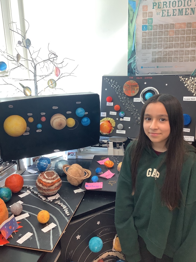 Young girl stands smiling beside a 3D solar system model featuring labeled planets and a periodic table poster, creating an educational atmosphere.