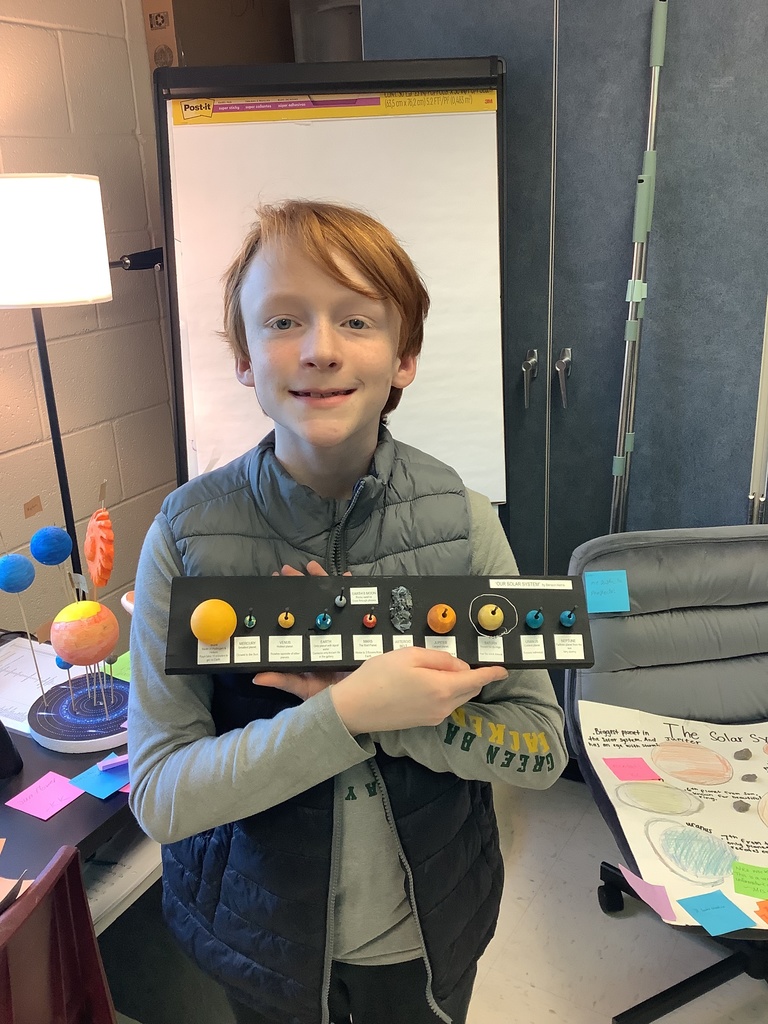 A young person with red hair smiles while holding a small model of the solar system. In the classroom background, there's a poster and a lamp. The atmosphere is educational and enthusiastic.