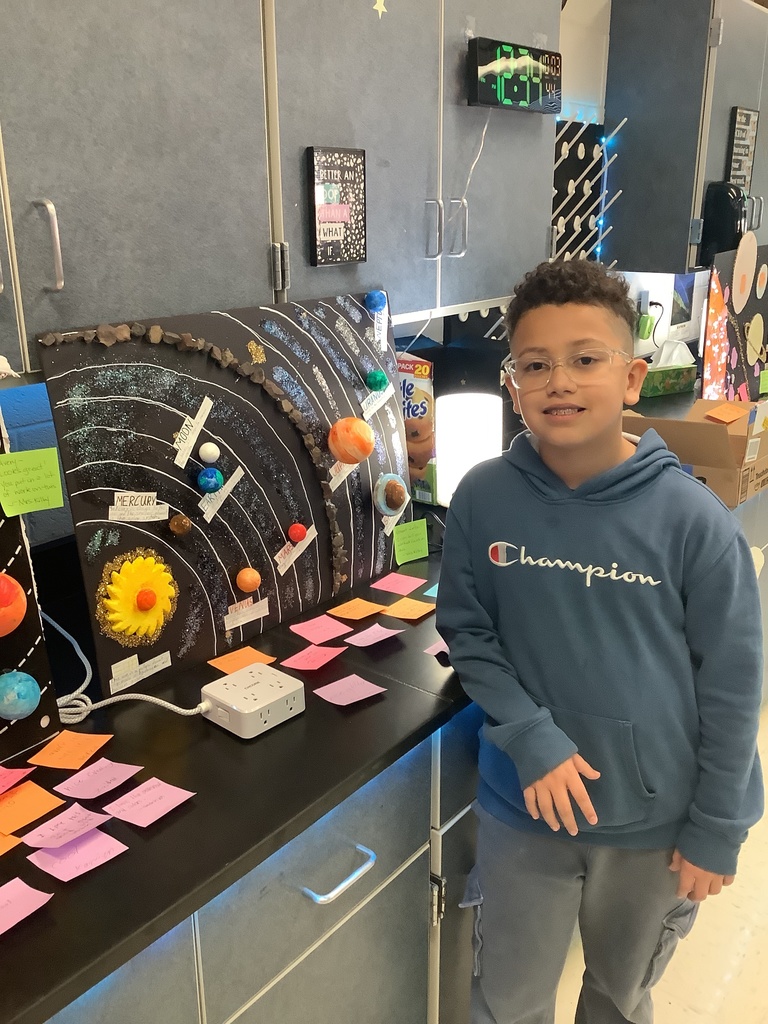 A young boy stands proudly beside his solar system project, featuring labeled planets on a black background. Bright post-it notes cover the table.