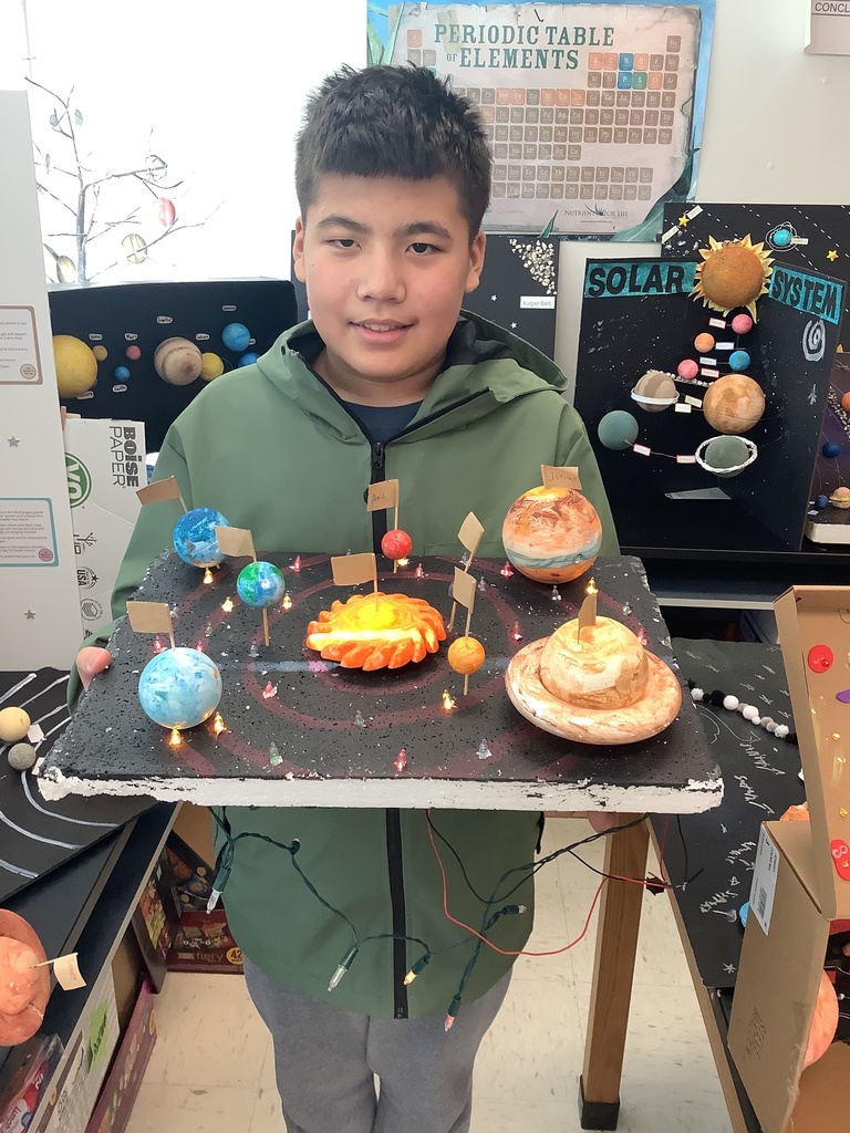 A young person holds a colorful solar system model with labeled planets, displayed under classroom posters. They are smiling, conveying pride and accomplishment.