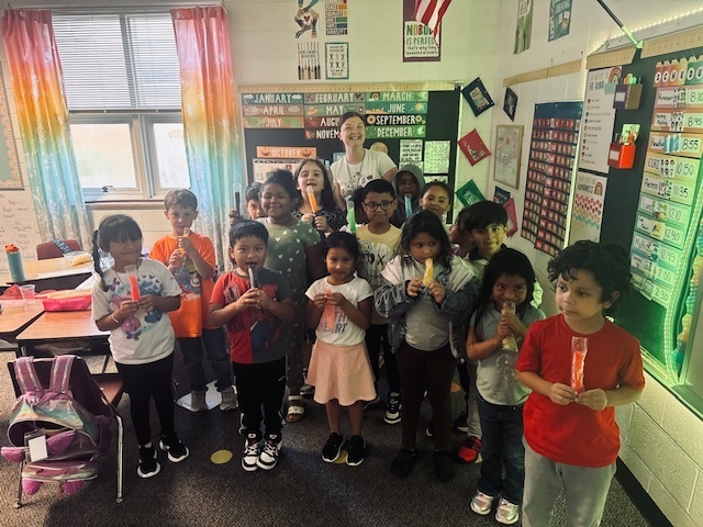 A classroom of young children smiling and holding popsicles, with a teacher standing in the back. The students are gathered together near colorful curtains and classroom decorations.
