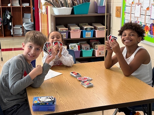 Three children sit at a classroom table smiling and holding up UNO cards. A deck of cards sits on the table. Shelves of books and bins are visible in the background.