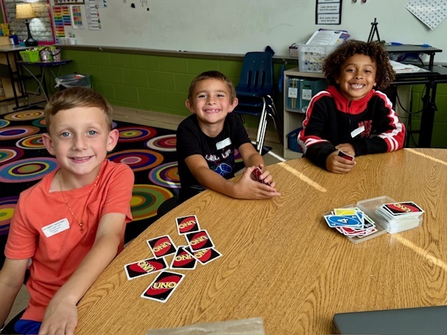 Three boys sit at a round classroom table playing UNO. They smile toward the camera with cards spread in front of them. A colorful rug and a whiteboard are visible behind them.