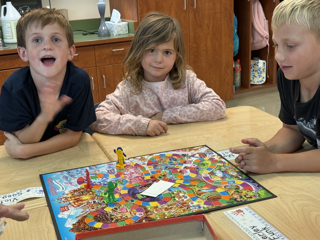 Three children sit at a classroom table playing Candy Land. They look at the colorful board game and pieces while smiling and talking together.