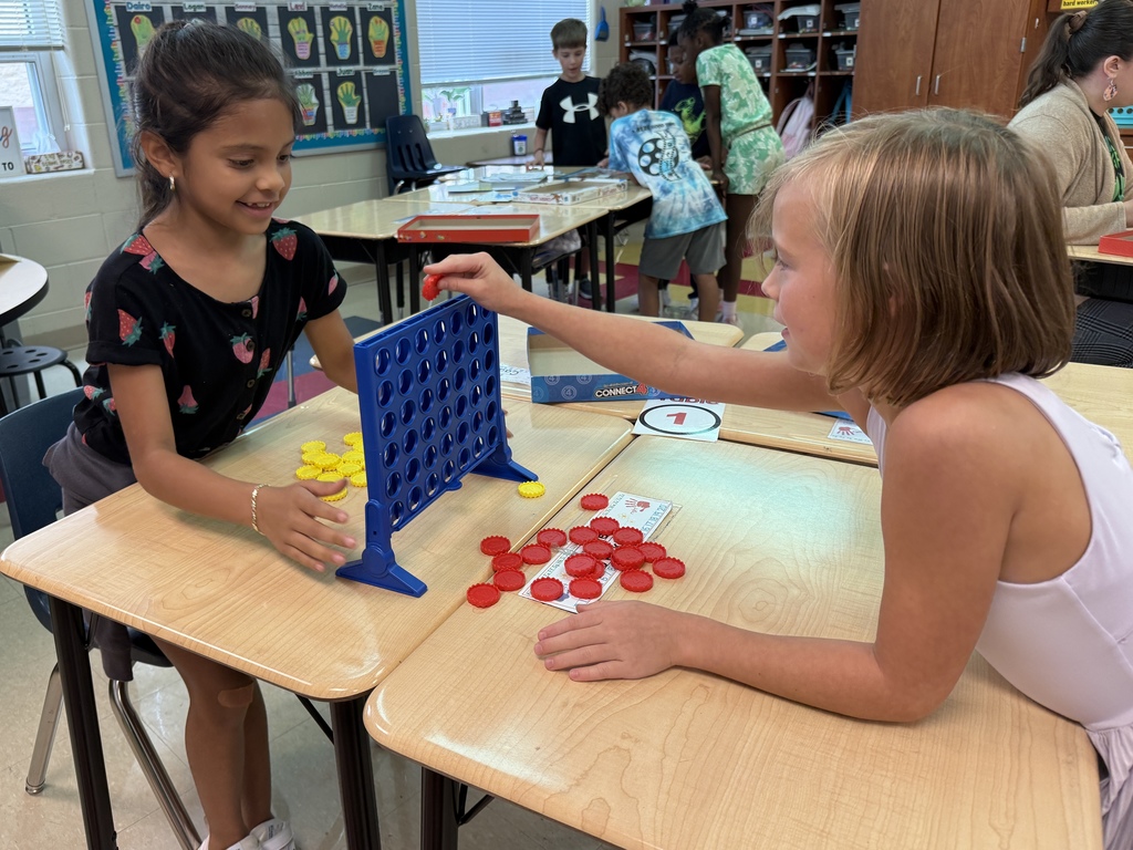 Two girls play Connect Four at a classroom desk. One girl places a red checker into the blue game board while both smile and focus on the game.