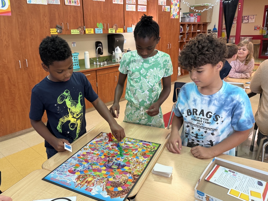 Three students stand around a Candy Land board game, concentrating as they move their game pieces. Other students can be seen working at tables in the background.