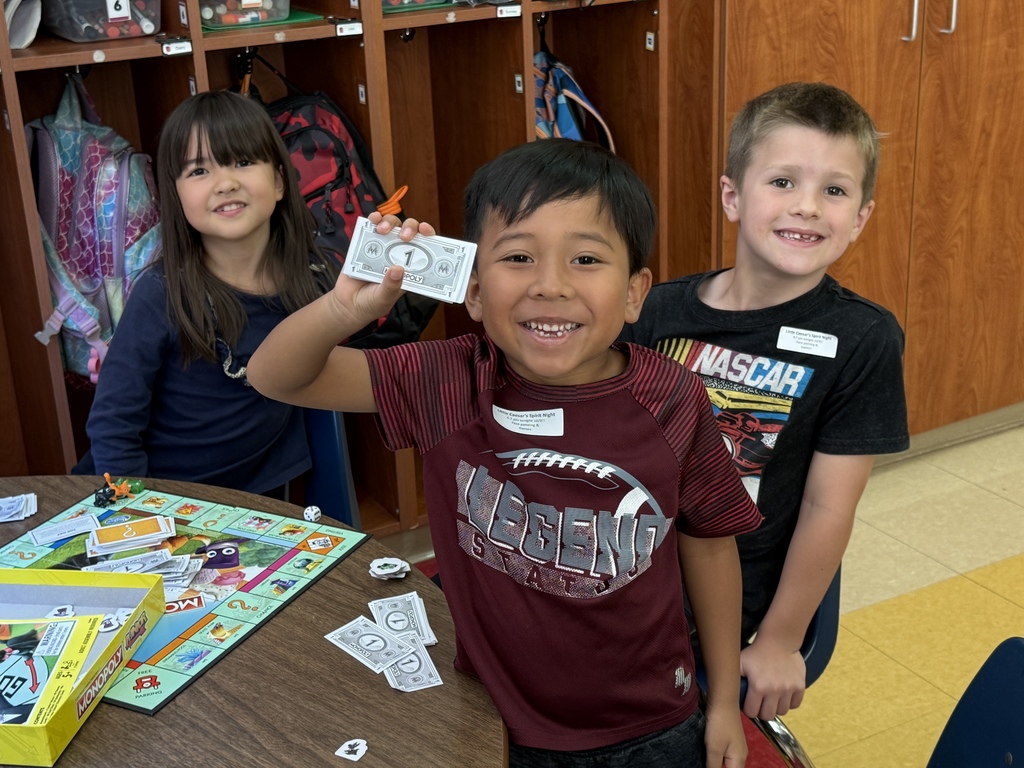 Three children stand around a table playing Monopoly Junior. One boy in a maroon shirt smiles widely while holding up play money, with game pieces and bills scattered on the table.