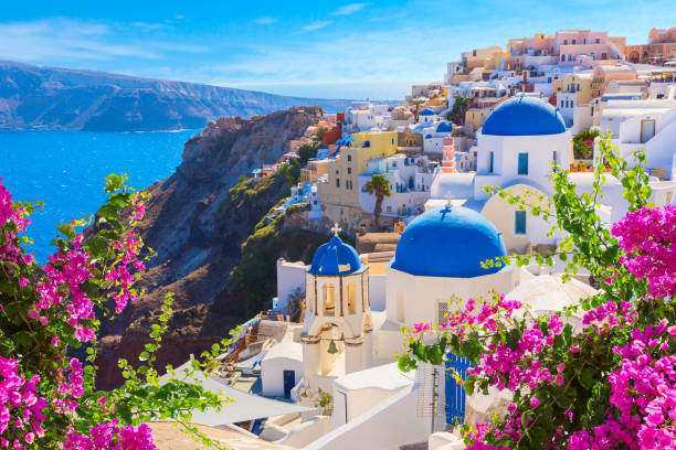 A breathtaking view of Santorini, Greece, showing whitewashed buildings with blue-domed roofs perched on a steep cliff overlooking the deep blue Aegean Sea. Bright pink bougainvillea flowers frame the foreground, while sunlight illuminates the hillside village cascading down toward the water.