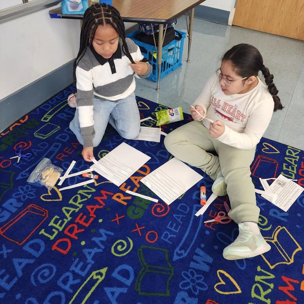Two students on the carpet cutting and gluing paper strips for a worksheet activity.