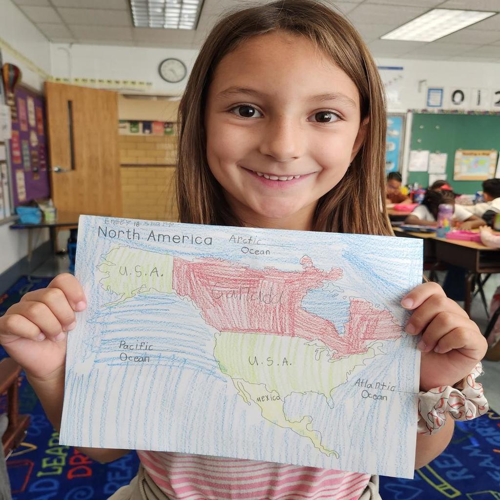 A student proudly holding a colored map of North America in a classroom.