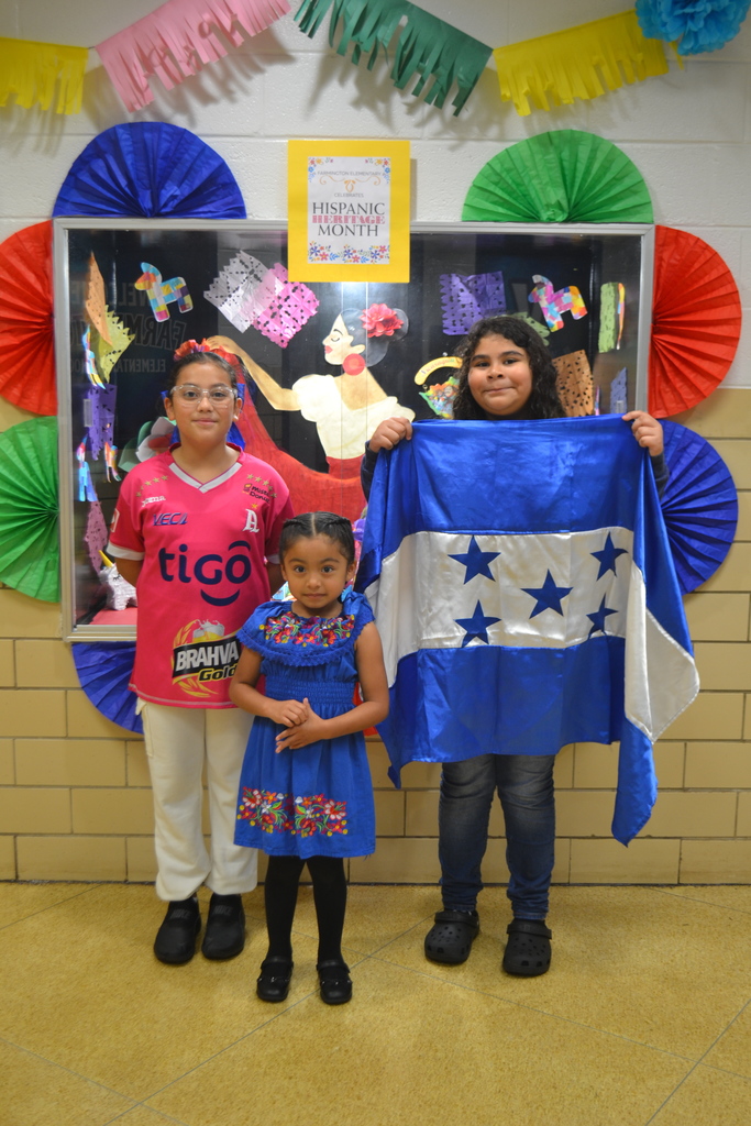 Three students in Hispanic Heritage spirit day clothing pose in front of the showcase decorated with bright colors and papel picado.