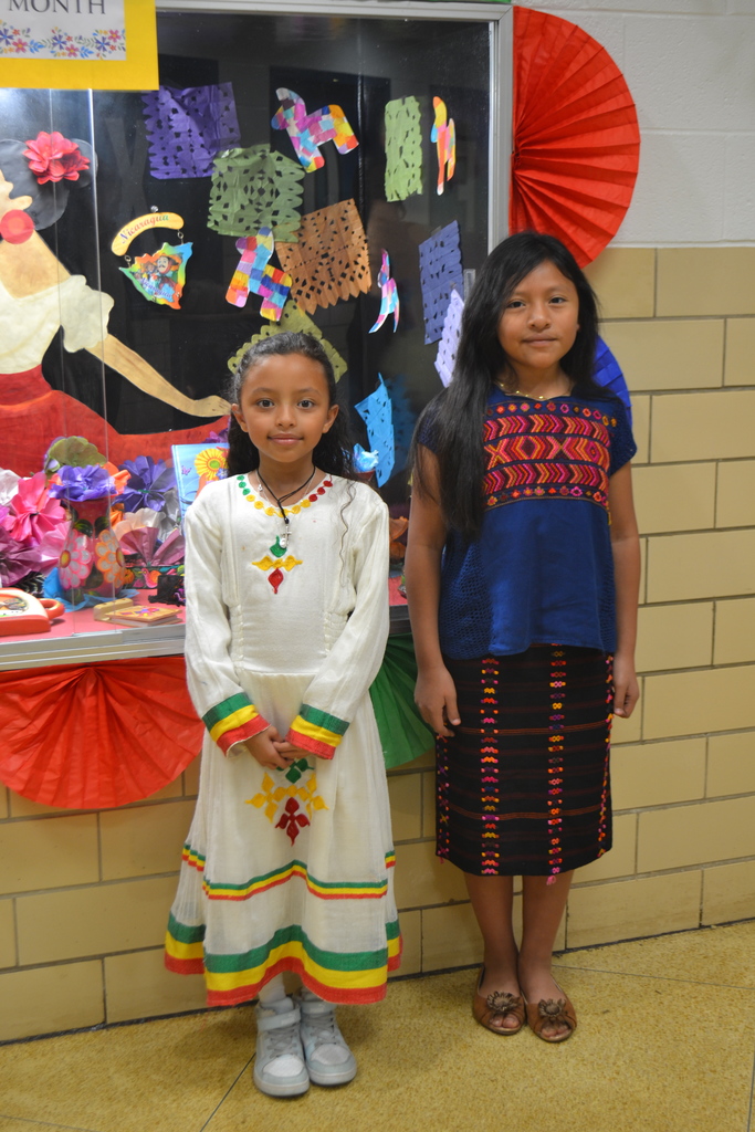 Two students in Hispanic Heritage spirit day clothing pose in front of the showcase decorated with bright colors and papel picado.