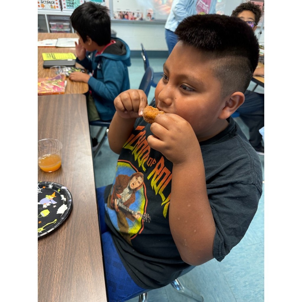 A student in a black “School of Rock” shirt eating a donut at a classroom table, with another student visible in the background.