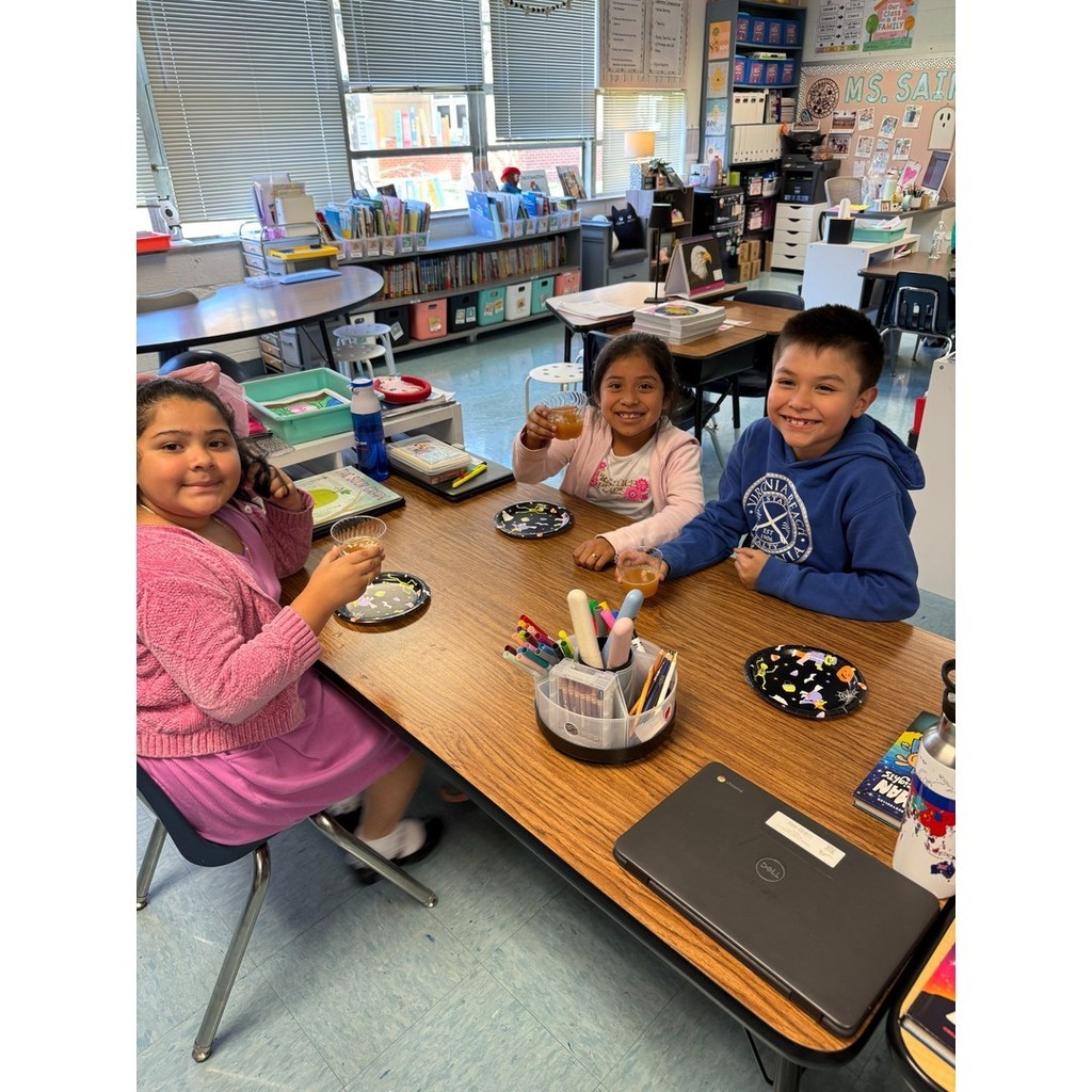 Three smiling students sitting together at a classroom table with Halloween plates and cups. Bookshelves and bins are visible in the background.