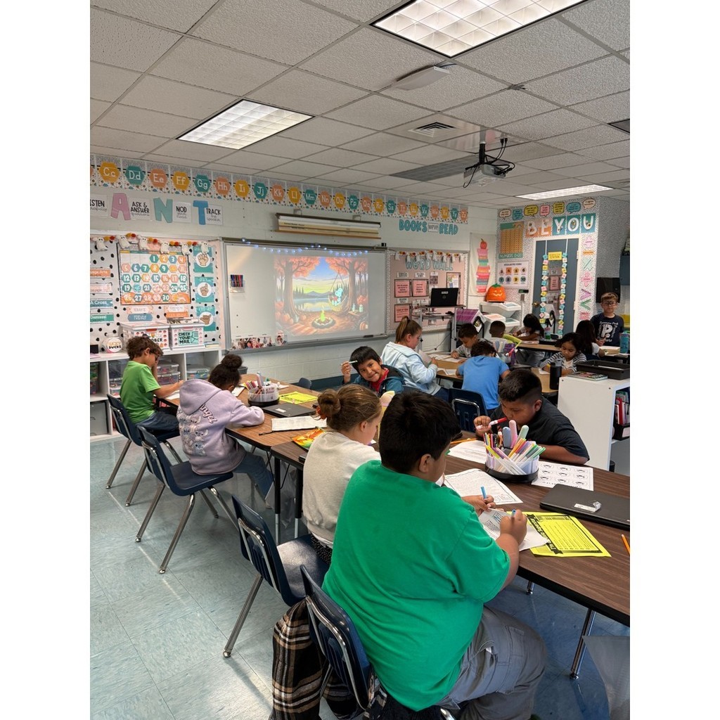 A classroom full of students working at tables with notebooks and papers. A projector shows a colorful fall forest scene on the whiteboard.
