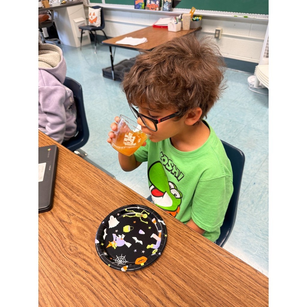 A student in a bright green Yoshi shirt sipping orange-colored punch from a clear cup. A black Halloween-themed plate sits on the desk.