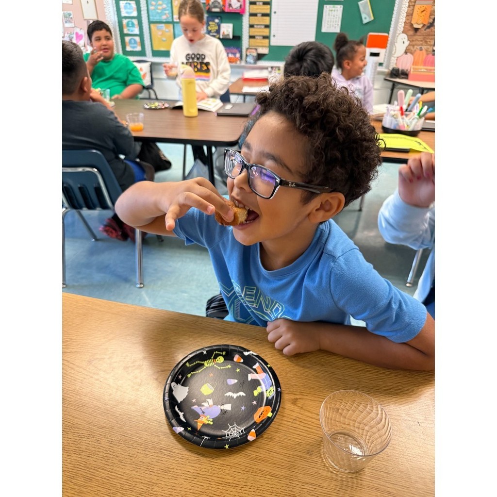 A student in glasses and a blue shirt smiling while eating a donut. Other students can be seen working and talking in the background.