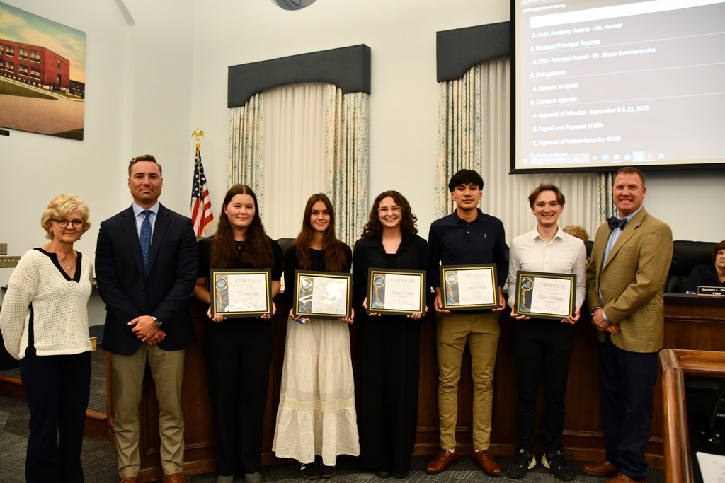 Five students holdnig certificates in a board room with a board member and principal on the left and Principal on the right