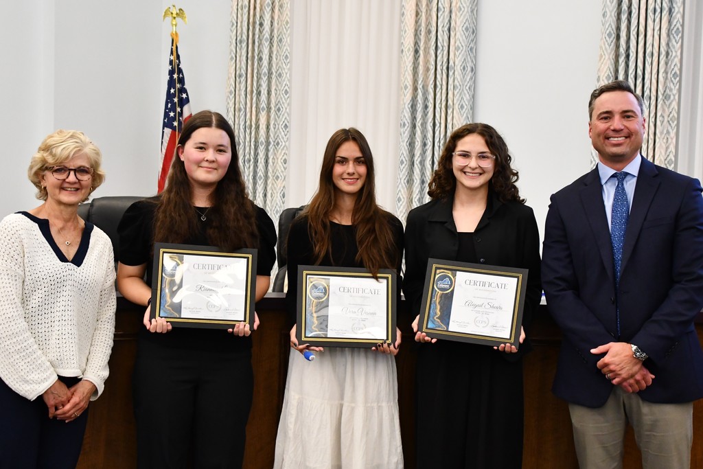three students holding certificates in a board room with a board member on the left and principal on the right