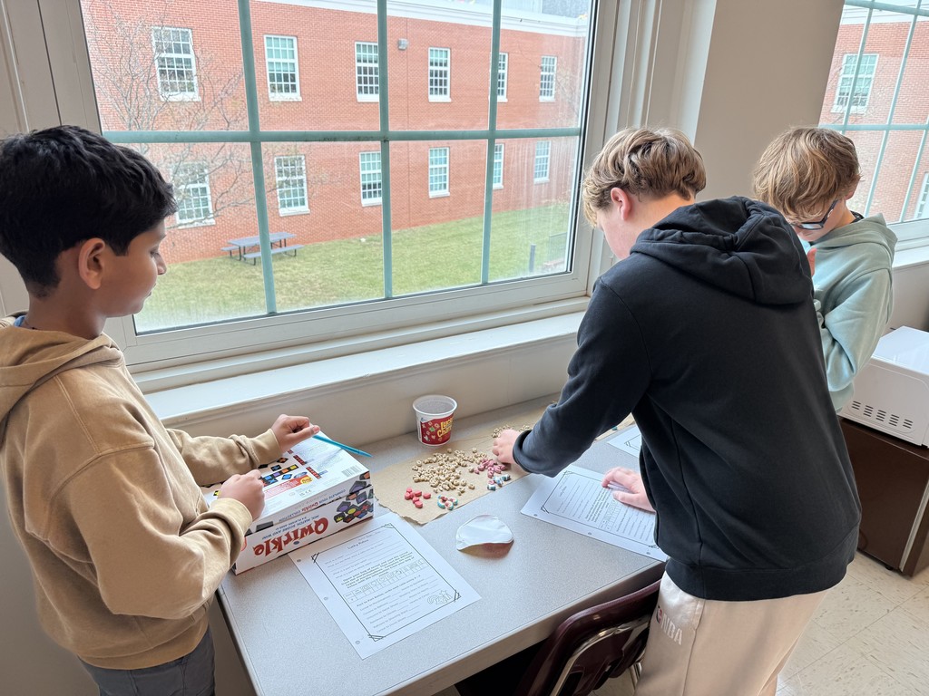Three students engaging in a classroom activity by a window, sorting cereal and candy on a table. Papers are spread out, creating a focused, collaborative atmosphere.
