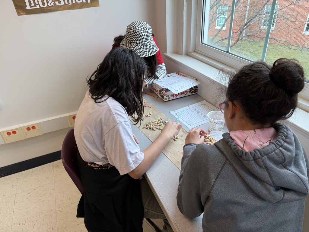 Three children sit at a table by a window, engaged in a craft activity with cereal and paper. The scene is warm and collaborative.