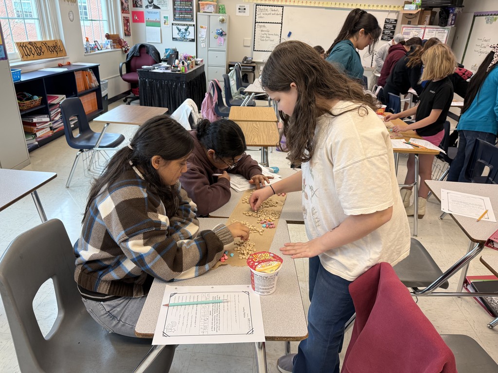 Students in a classroom sort colorful cereal loops on a table, engaged and focused. Papers and a cereal box are scattered around, creating a lively educational atmosphere.
