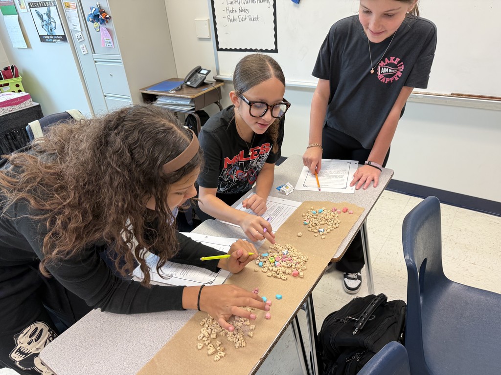 Three middle school students work together at a classroom table covered with a strip of brown paper and piles of cereal pieces. They are smiling and using pencils to sort or count the pieces as part of a hands-on classroom activity. Worksheets and school supplies are on the table around them.