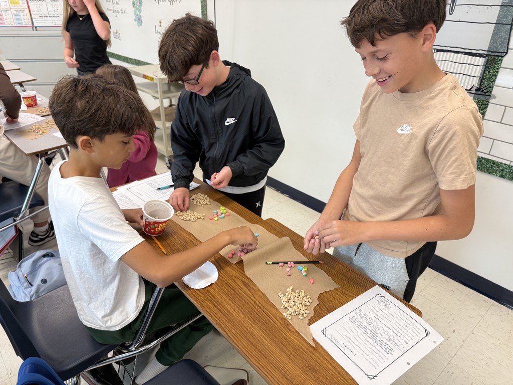 Three boys are smiling and sorting cereal pieces at a school desk. Papers and colored cereal are scattered as they engage in a fun classroom activity.