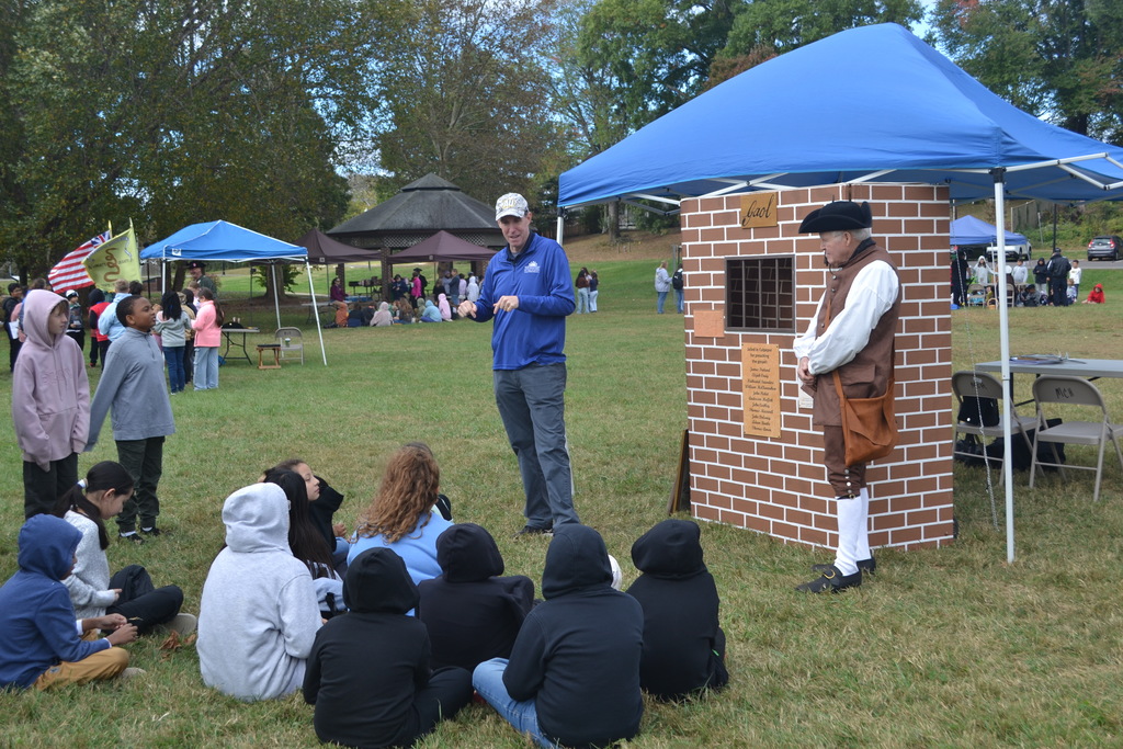 Man in blue shirt presents next to a colonial dressed man in the park to a group of students.