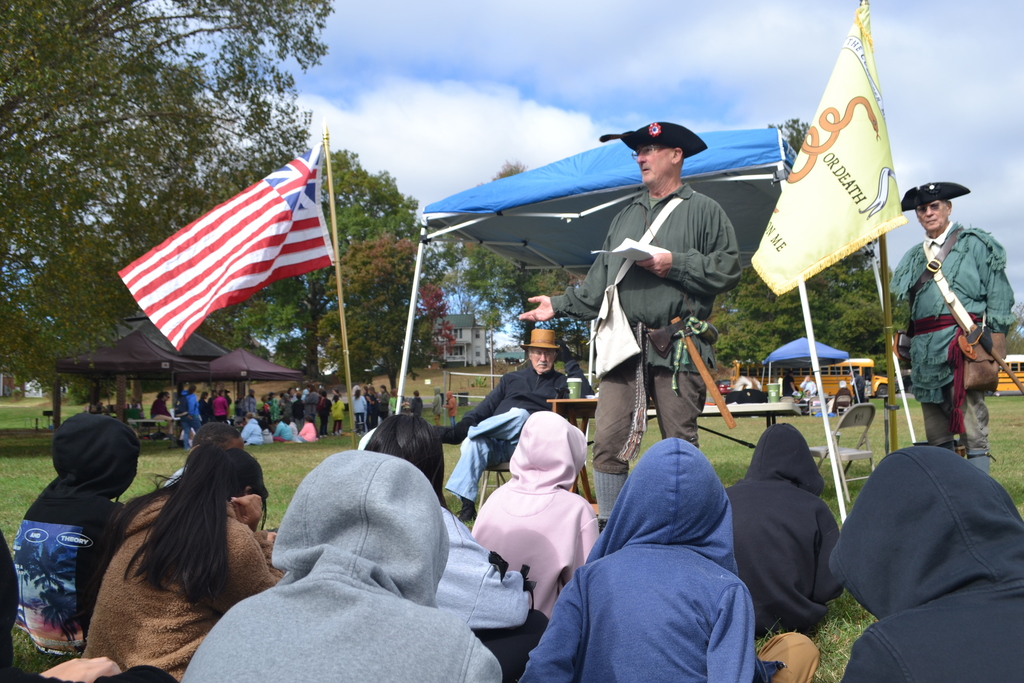 Minute men dressed reinactors present before a group of kids in the park.  Flags flying in th background.