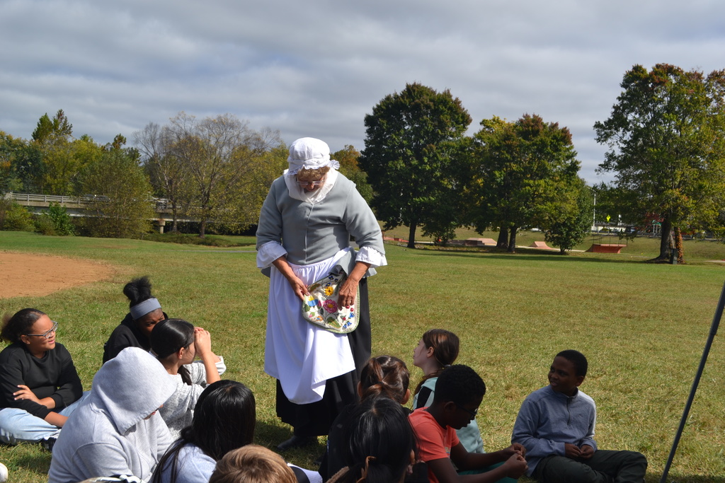 Lady in colonial clothing shows her pocket purse to a group of students in a park.