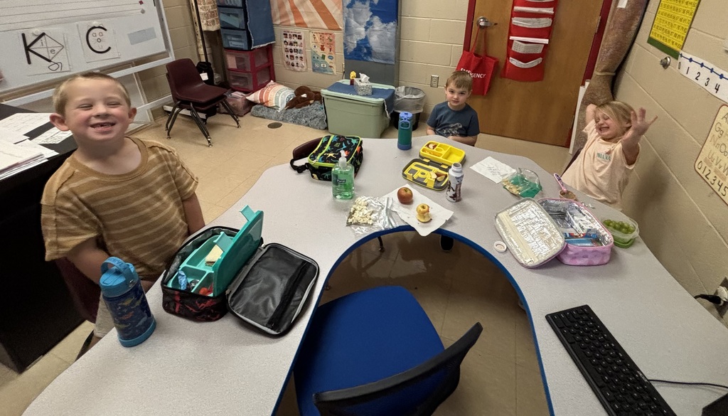 Three children sit at a curved classroom table enjoying lunch. They have lunchboxes, drinks, and snacks in front of them and are smiling and waving at the camera.