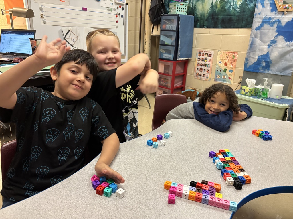 Three children sit together at a classroom table, smiling and waving. They are working with colorful linking cubes to build shapes and structures. The classroom background shows posters, storage bins, and learning materials.