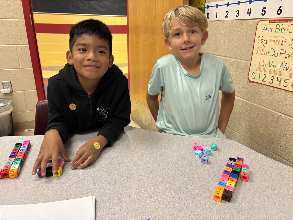 Two young students sit at a classroom table smiling at the camera. They are using colorful linking cubes to create towers and patterns. A number line and alphabet chart are visible on the wall behind them.