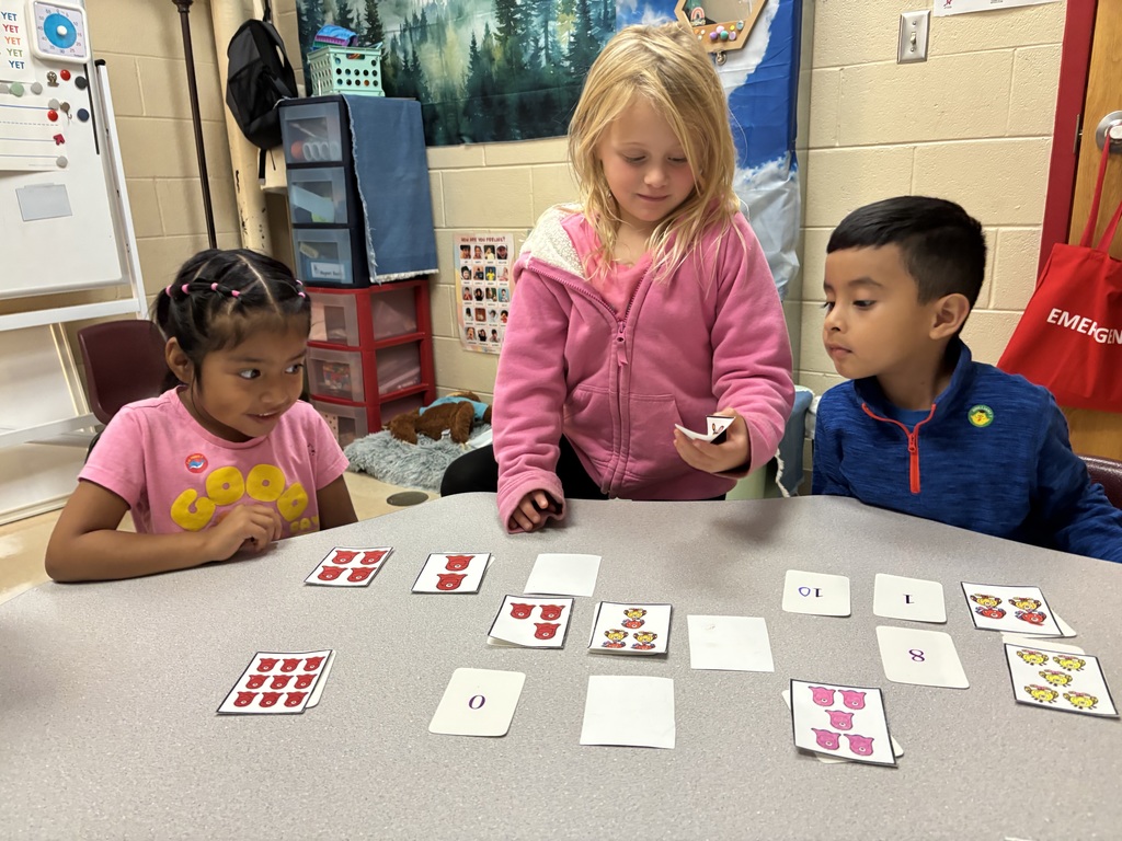 Three children sit at a classroom table sorting and matching picture and number cards. One child is holding up a card while the others watch.
