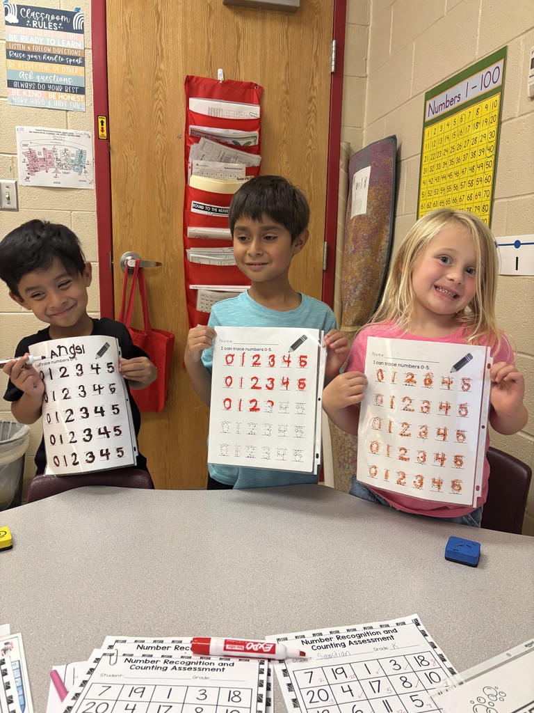 Three children proudly hold up their worksheets showing traced numbers 0 through 5. They are smiling and standing in front of a classroom door with learning posters on the wall.