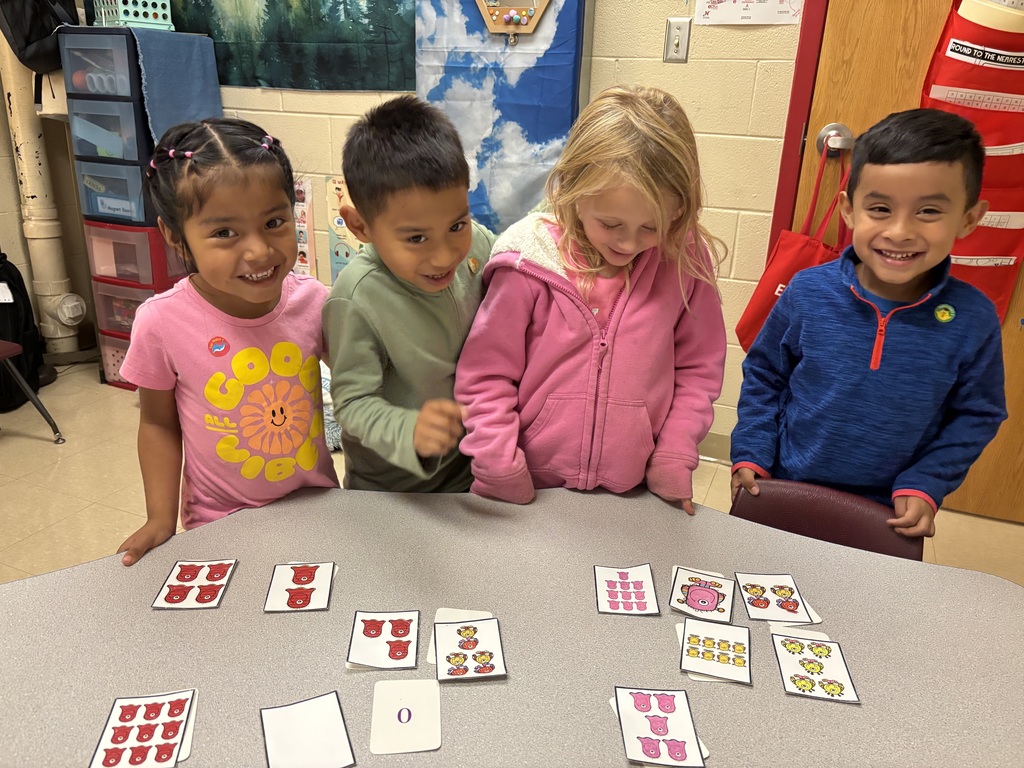 Four children stand around a classroom table looking at number and picture cards showing groups of objects. They are smiling and engaged in a counting activity.