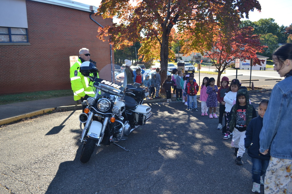 A class is looking at the motorcycle and the police officer outside of the school on a sunny fall morning.