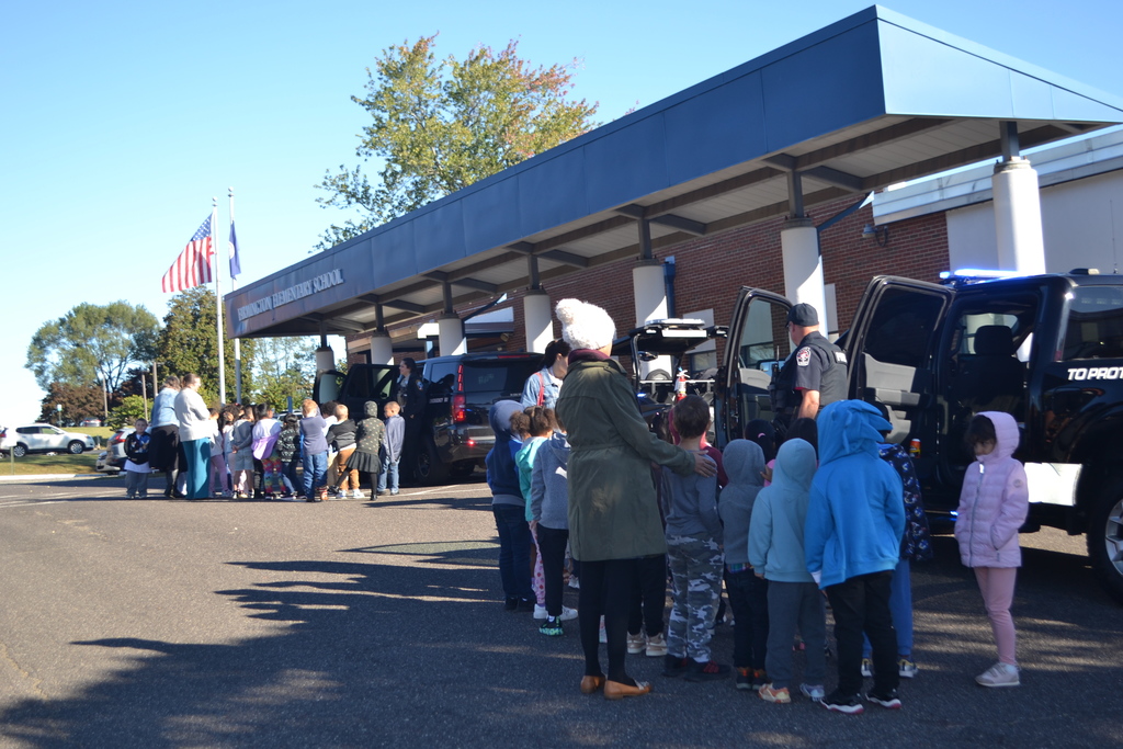 Two classes are outside of the school (Farmington Elementary) Each class is looking and talking with a police officer.