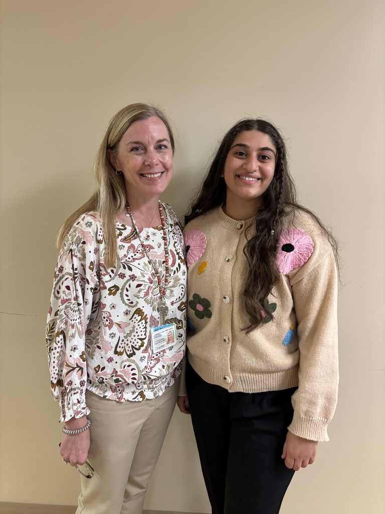 Two people stand smiling side by side in front of a beige wall. The person on the left has long blonde hair and wears a light floral blouse with tan pants, holding glasses in one hand and wearing a school ID badge on a lanyard. The person on the right has long dark hair and wears a tan cardigan decorated with colorful flower designs and black pants.