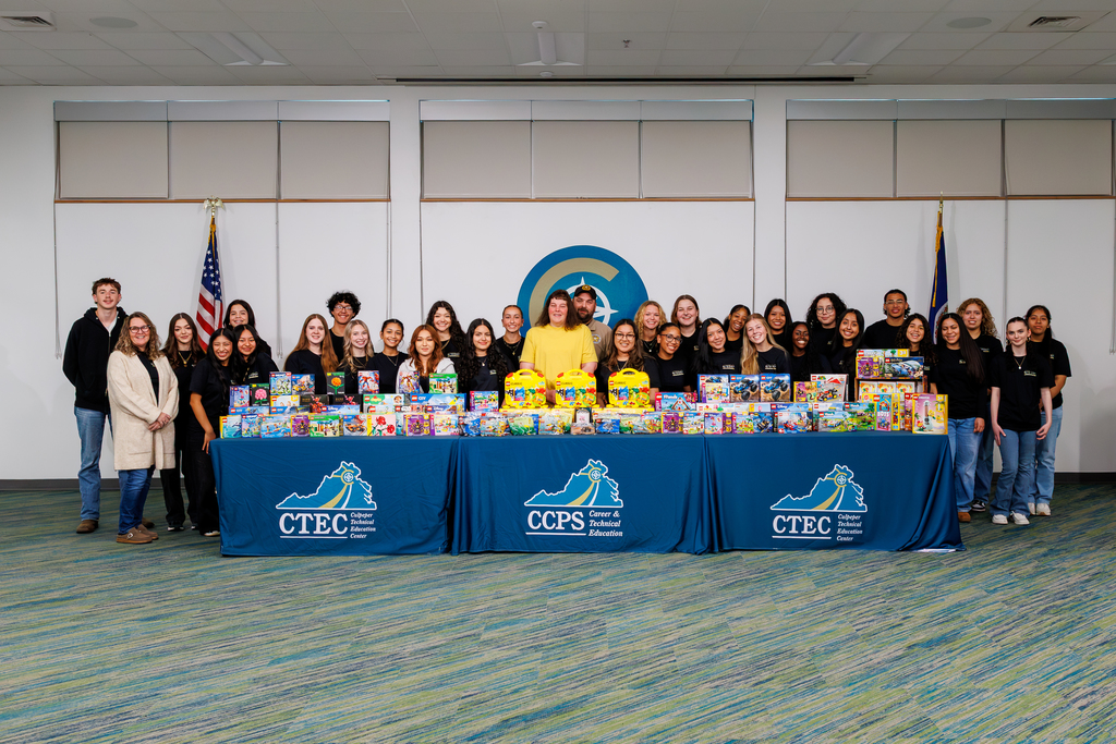 CTEC HOSA students and staff stand together behind tables filled with LEGO sets collected during a LEGO drive to support children battling childhood cancer. The tables are covered with Culpeper County Public Schools and CTEC banners, and everyone smiles proudly in front of the CTEC logo backdrop and U.S. flags.