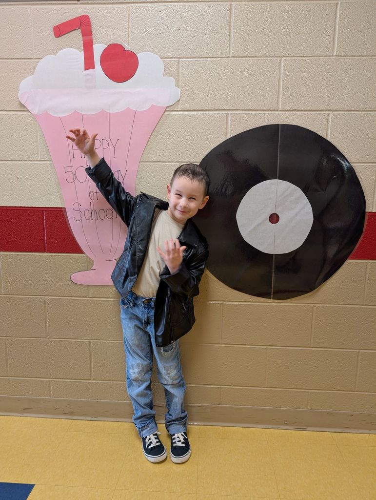 A young student poses in front of the “Happy 50th Day of School” wall display wearing a leather jacket, jeans, and sneakers. He makes a rock ’n’ roll hand gesture and smiles proudly.