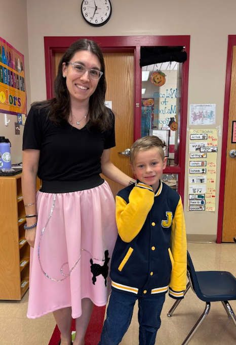 A teacher wearing a black top and pink poodle skirt smiles beside a student in a letterman jacket with yellow sleeves. They stand inside a classroom with colorful posters and learning materials in the background.
