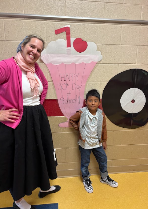A teacher wearing a pink cardigan, scarf, and black poodle skirt smiles beside a young student dressed in a vintage-style shirt and checkered shoes. Behind them is a large paper milkshake decoration with text that reads “Happy 50th Day of School!” and a paper record on the wall.