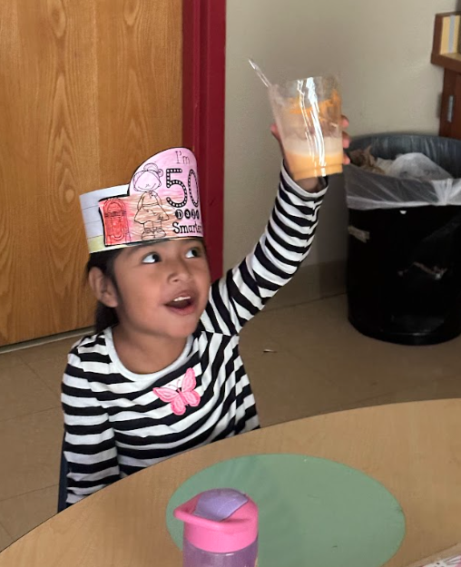 A young student wearing a striped shirt and a handmade paper hat that says “I’m 50 Days Smarter” raises her cup in celebration. She is smiling while enjoying a root beer float at her classroom table.