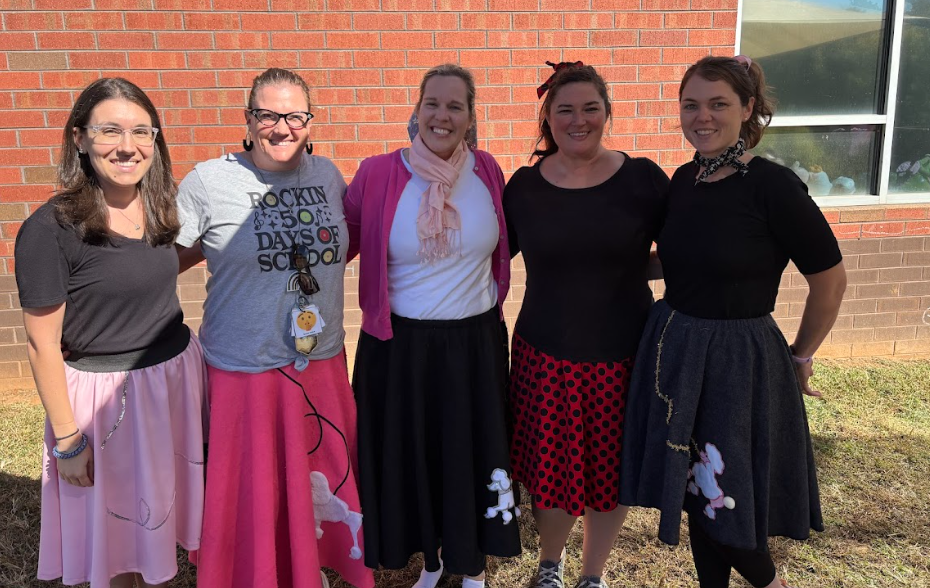 Five teachers stand smiling outside in front of a brick wall, dressed in 1950s-themed outfits. They are wearing poodle skirts, scarves, and glasses, with one shirt reading “Rockin’ 50 Days of School.” Sunlight shines on the group as they pose together.