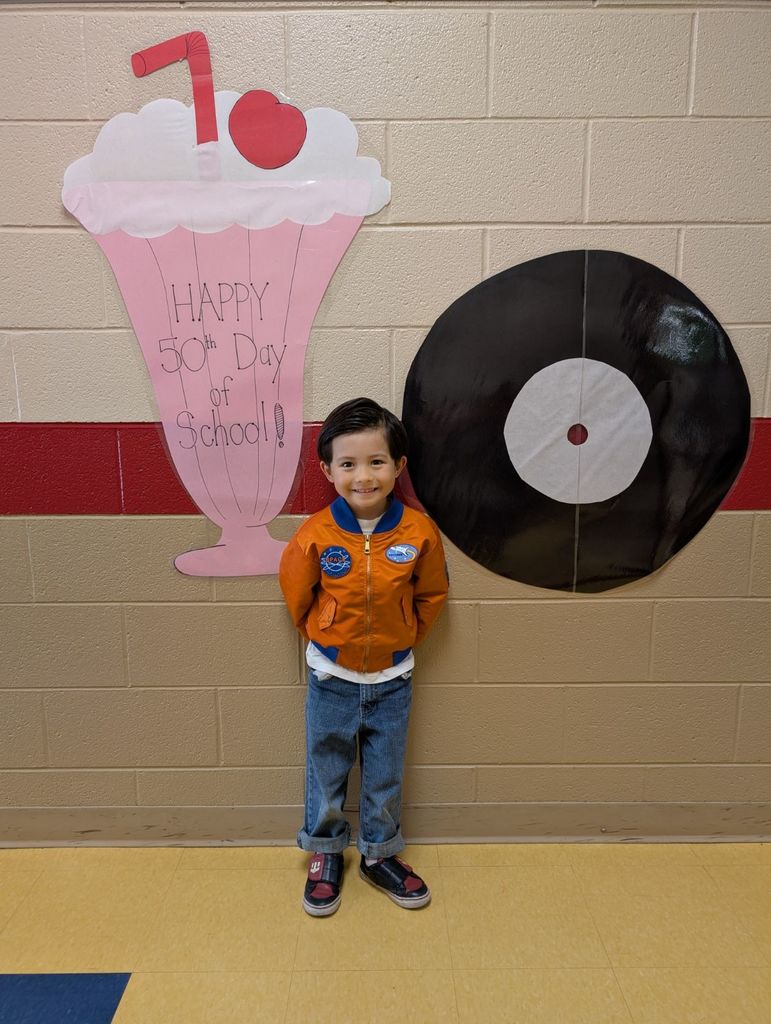 A student stands in front of the same “Happy 50th Day of School” wall display wearing a bright orange jacket with NASA patches, rolled-up jeans, and sneakers. He smiles happily for the photo.