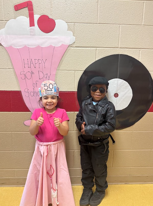 Two students stand smiling in front of a wall decorated with a large paper milkshake and record that say “Happy 50th Day of School.” One child wears a pink poodle skirt and “50 Days Smarter” hat, while the other is dressed in a black leather jacket and sunglasses.