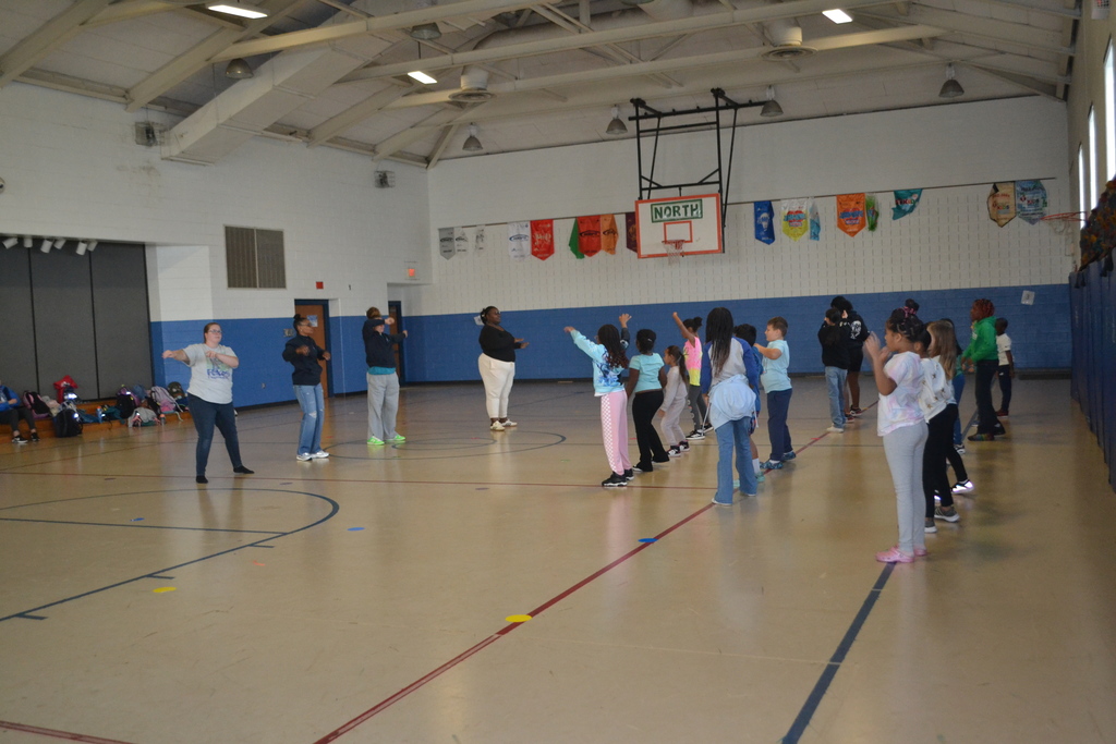 Image contains 3 rows of students dancing in a line and 4 adults leading them in the school gym.