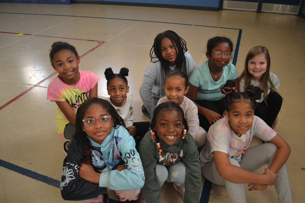 Image contains a group of 9 girls smiling for a picture during their club in the school gym.